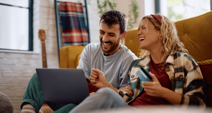 Szczęśliwa para siedzi na podłodze, przeglądając promocje na laptopie i trzymając kartę podarunkową, co pokazuje, co motywuje klientów do zakupu.