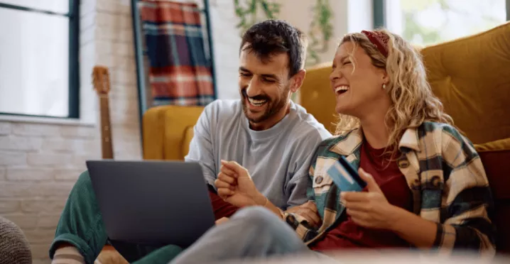 Szczęśliwa para siedzi na podłodze, przeglądając promocje na laptopie i trzymając kartę podarunkową, co pokazuje, co motywuje klientów do zakupu.