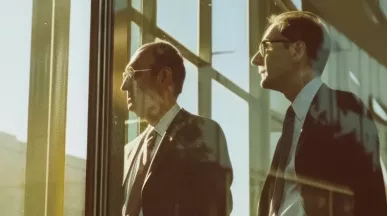 Dwaj mężczyźni w garniturach patrzący przez okno biurowca, oświetleni ciepłym światłem słońca